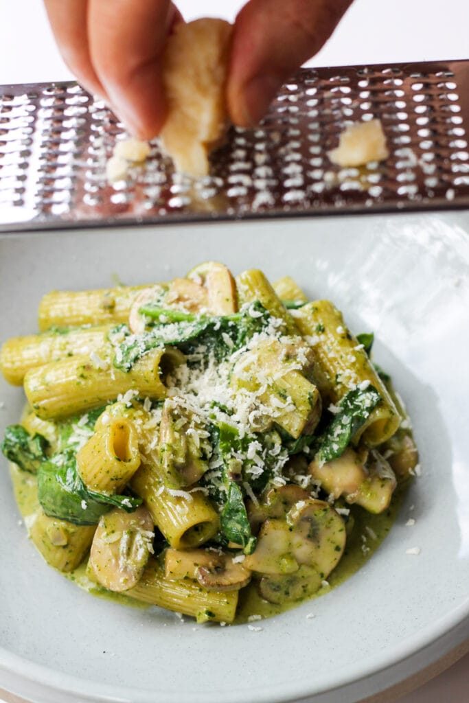 Grated parmesan over rigatoni with brown mushrooms and basil pesto sauce in a bowl