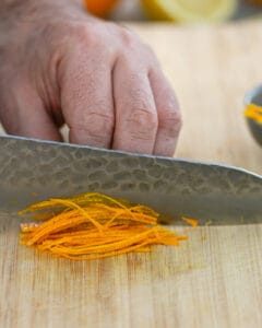 Picture showing how to julienne the orange zest with a sharp knife on a wooden chopping board