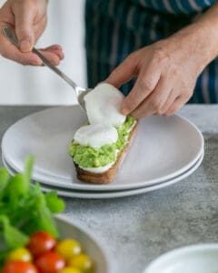 Placing two poached eggs on top of avocado mix, ricotta spread on toasted sourdough slice