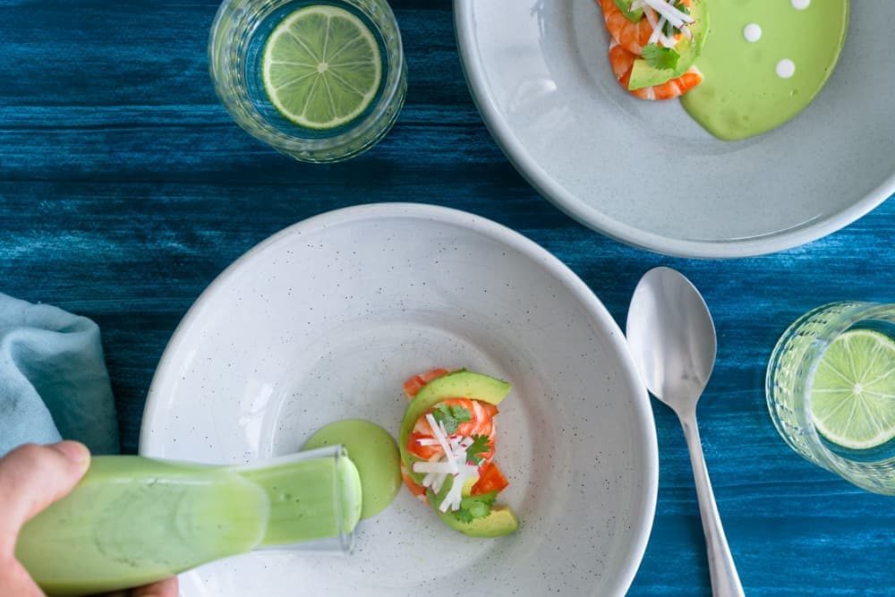 Overhead shot of prawn salad arranged in a bowl with avocado soup being poured from a glass jar