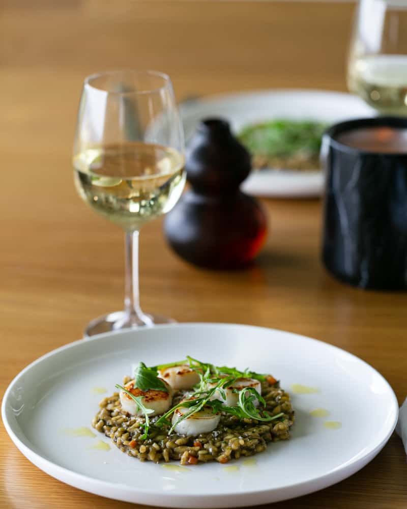 Risotto plated on a white plate topped with 4 scallops and arugula leaves with a glass of white wine