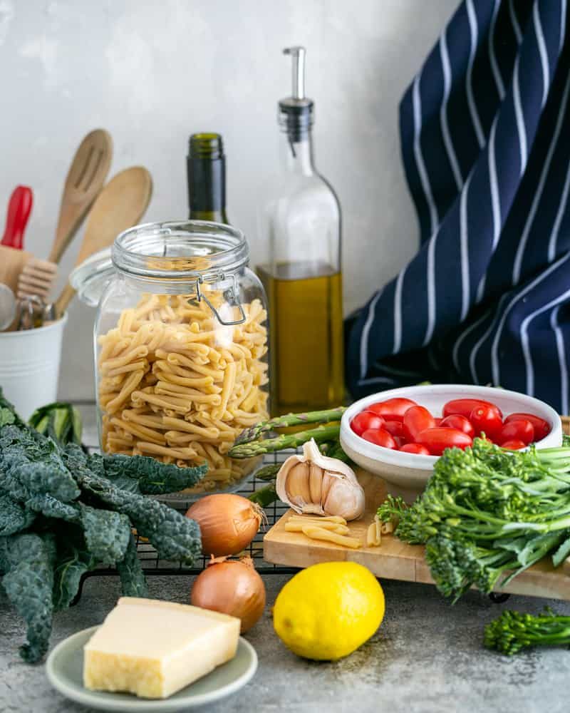 Picture with ingredients. Bowl of baby tomatoes, kale, asparagus, parmesan block, lemon, shallot, garlic, kale, pasta, olive oil.