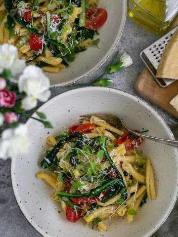 Two bowls of Casarecce Pasta with Asparagus, Kale and Garlic with Parmesan block on grater on a wooden board