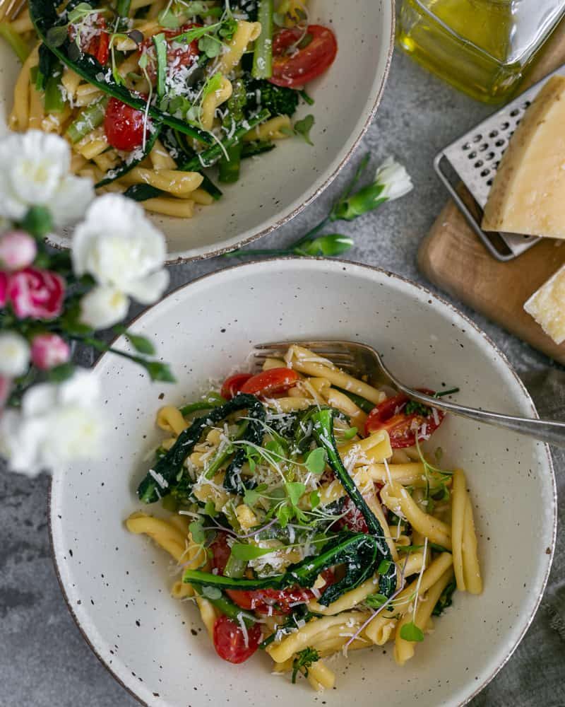 Two bowls of Casarecce Pasta with Asparagus, Kale and Garlic with Parmesan block on grater on a wooden board