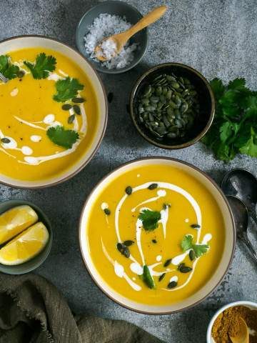 Two bowls of pumpkin soup garnished with pepitas, fresh coriander and coconut milk