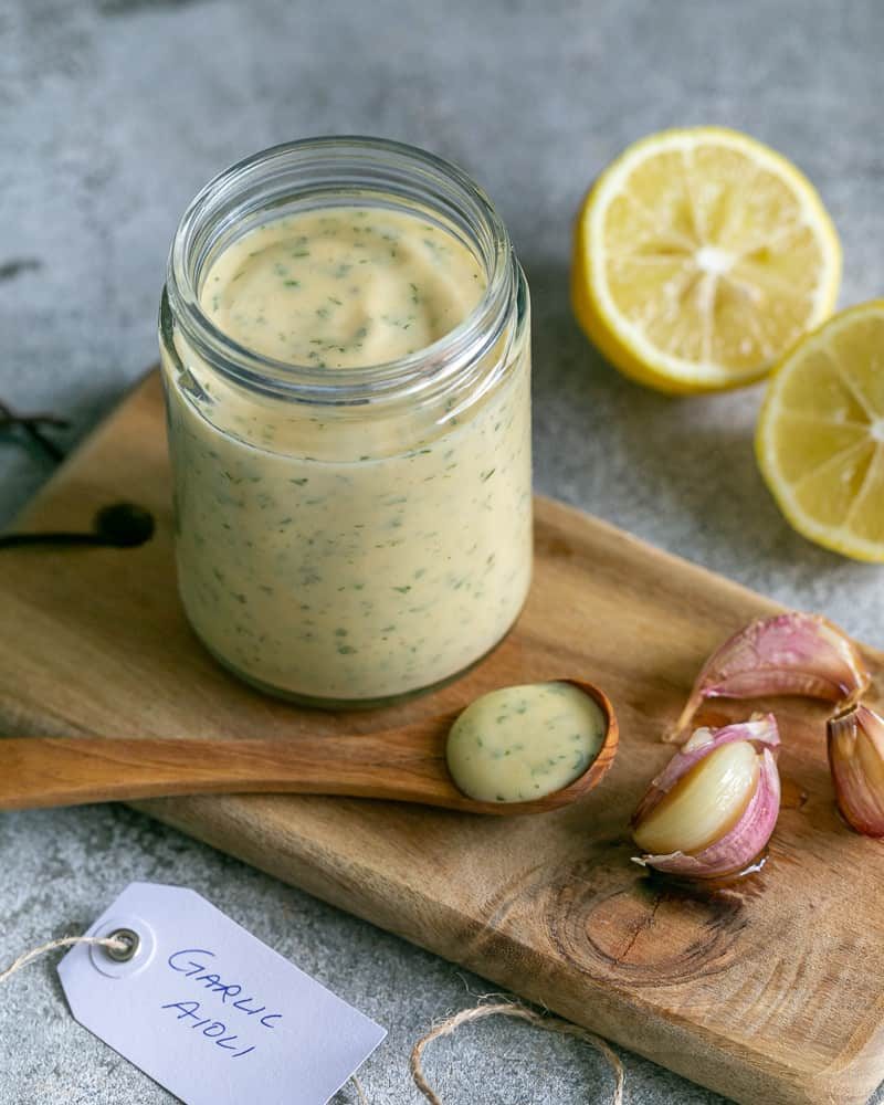 Garlic Aioli in a glass jar with a wooden spoonful of the aioli in front and on the side 3 confit garlic cloves and a lemon cut in half in the background