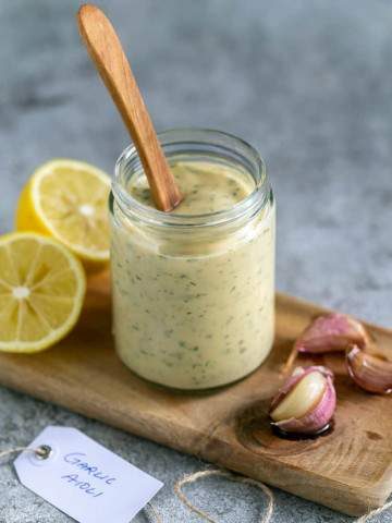 Garlic Aioli in a glass jar with a wooden spoon in it on the side 3 confit garlic cloves and a lemon cut in half in the background