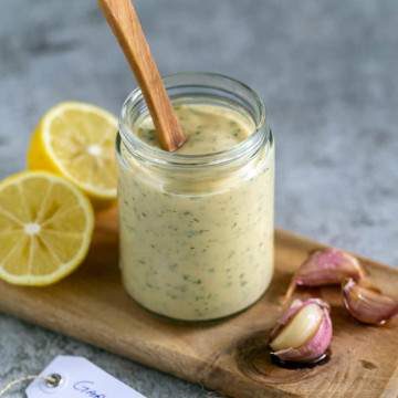 jar with confit garlic and herb aioli with a spoon inside. Lemon cut in half on the left and garlic cloves confit on the right.