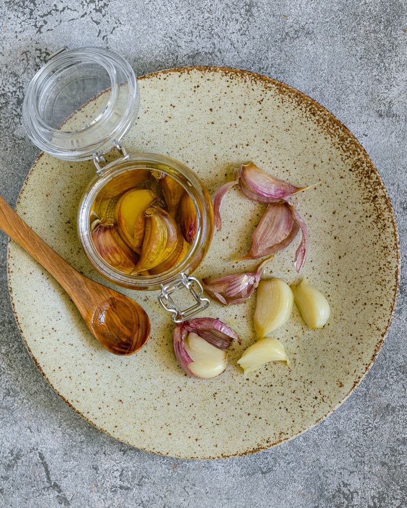 Confit garlic in a small glass jar on a small ceramic plate with a wooden spoon on the side to make aioli sauce