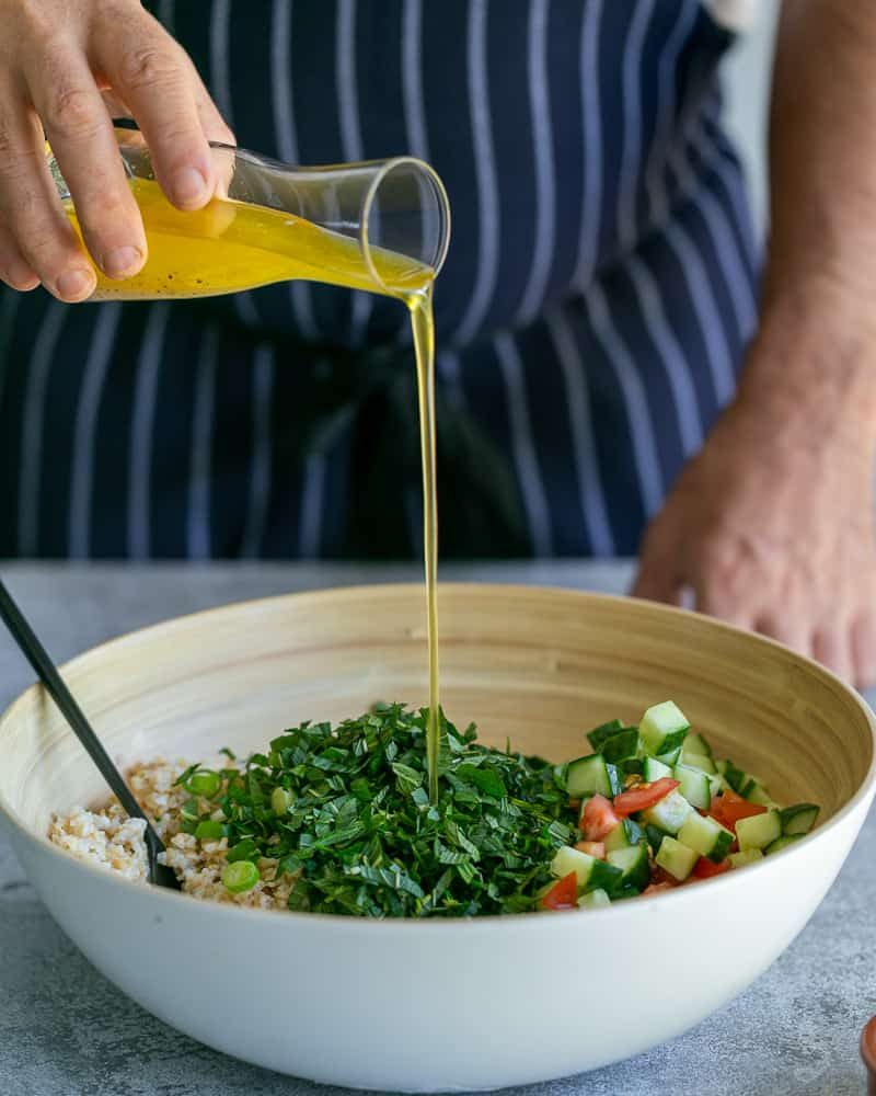 Adding salad dressing to bowl with chopped parsley, tomatoes and cucumbers, bulgar, mint and spring onions