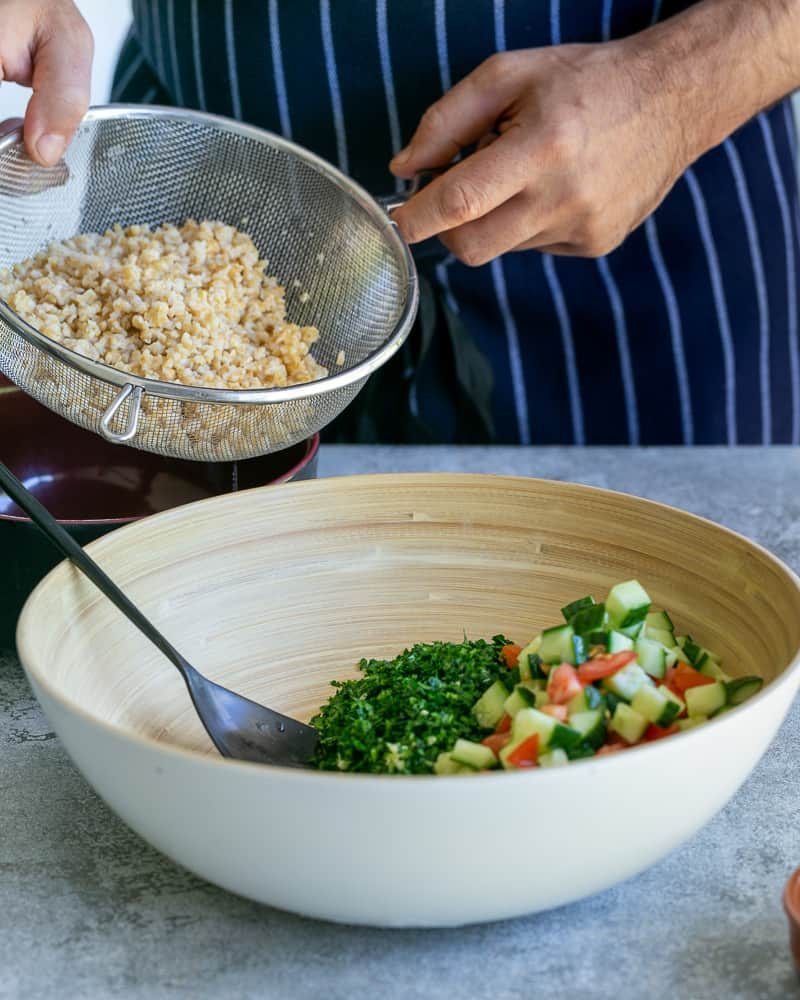 Adding cooked bulgar to bowl with parsley and chopped tomatoes and cucumbers