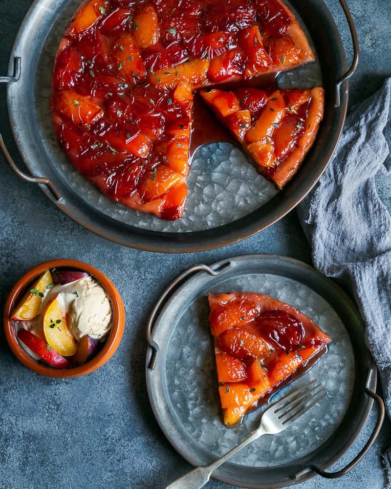 A slice of Plum Tarte Tatin on a vintage metal plate and a fork with the whole tarte tatin the background on a larger vintage metal plate served with macadamia ice cream and fresh sliced plums in a ceramic bowl