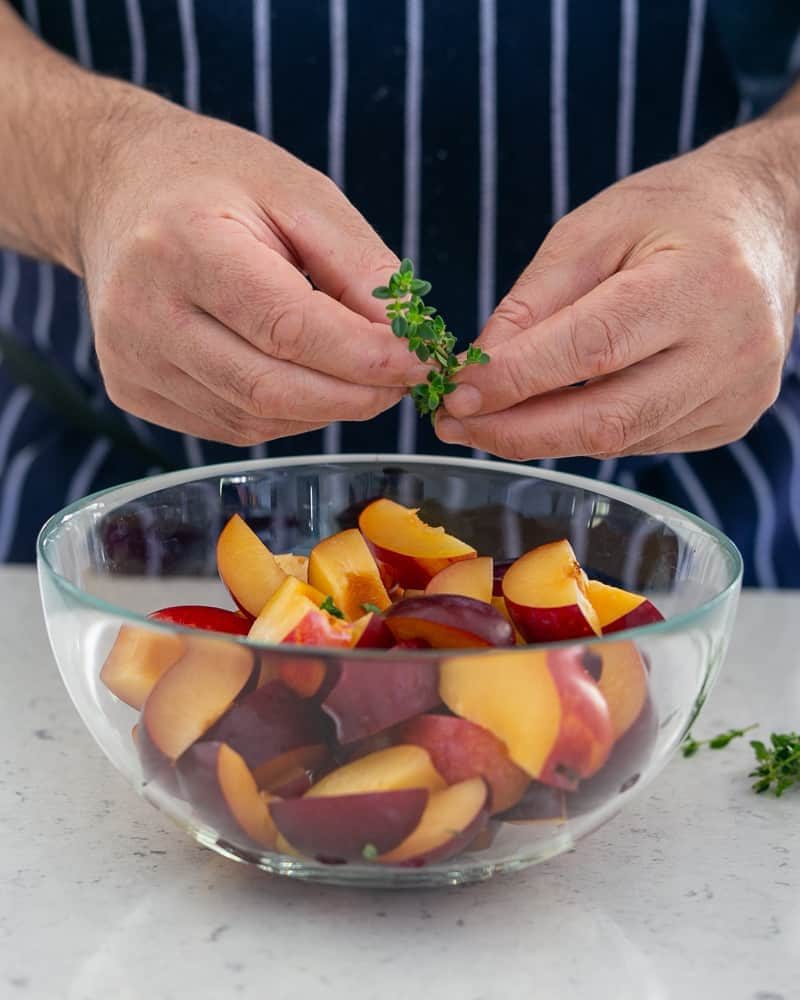 Adding a sprig of fresh lemon thyme to plum wedges in a glass bowl