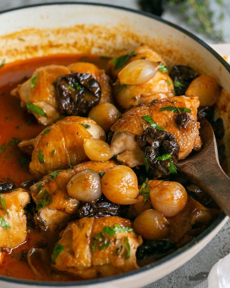 Chicken braised with pruned and shallots in a wide shallow pan with a wooden spoon scooping a chicken piece