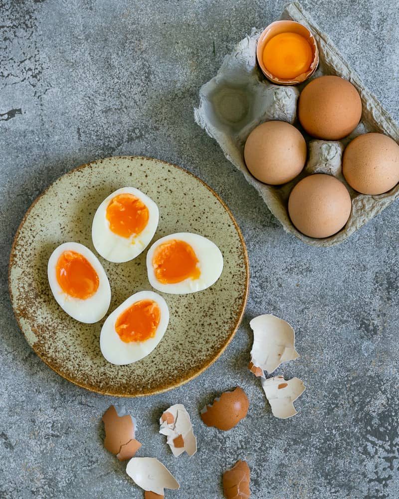 Soft boiled eggs in a ceramic plate showing the texture of the egg yolk. On the side a small tray of whole eggs and few broken egg shells from the peeled boiled eggs to make aioli sauce