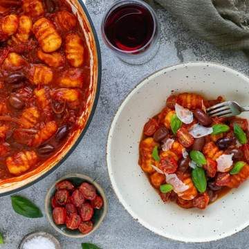 Ricotta Gnocchi's with Tomatoes and olives plated in a bowl with the Gnocchi pan in the background, oven dried tomatoes and shaved parmesan as garnishes with a glass of red wine on the side
