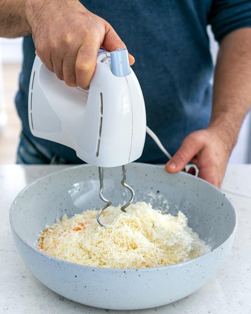 With a hand blender mixing parmesan cheese to eggs, flour and ricotta in a bowl 