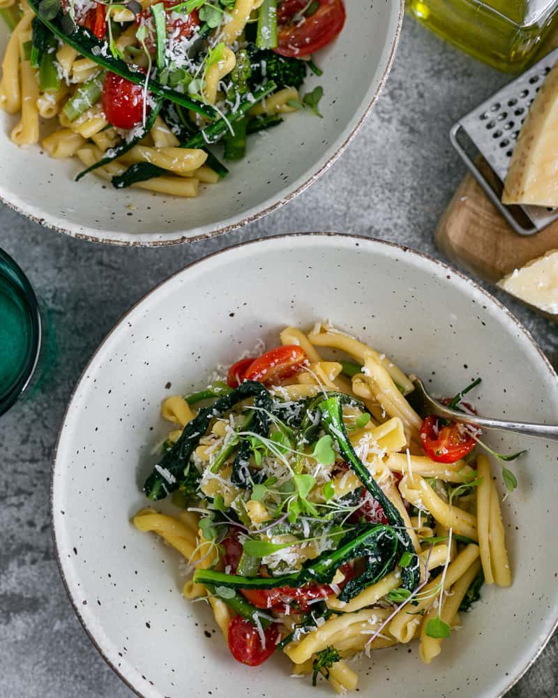 Two bowls of Casarecce Pasta with Asparagus, Kale and Garlic with Parmesan block on grater on a wooden board