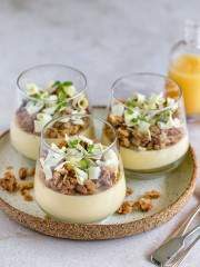 Lemon Yuzu Posset with white chocolate crumble in three verrine glasses on a round ceramic plate with three dessert spoons on the side and a bottle of Yuzu in the background