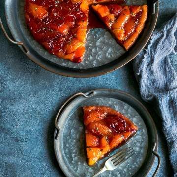 A slice of Plum Tarte Tatin on a vintage metal plate and a fork with the whole tarte tatin the background on a larger vintage metal plate