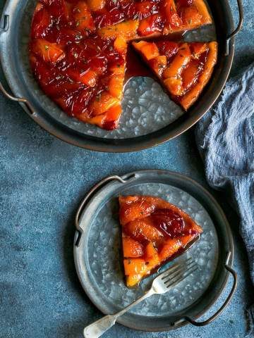 A slice of Plum Tarte Tatin on a vintage metal plate and a fork with the whole tarte tatin the background on a larger vintage metal plate