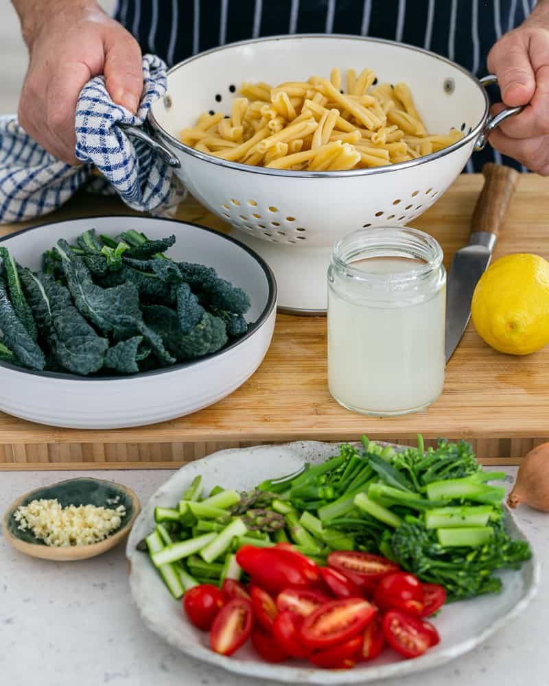 Freshly cooked Casarecce pasta in a strainer with kale in a bowl, pasta water in a jar, lemon, chopped garlic in a pinch bowl, chopped broccolini, asparagus and tomatoes in a plate
