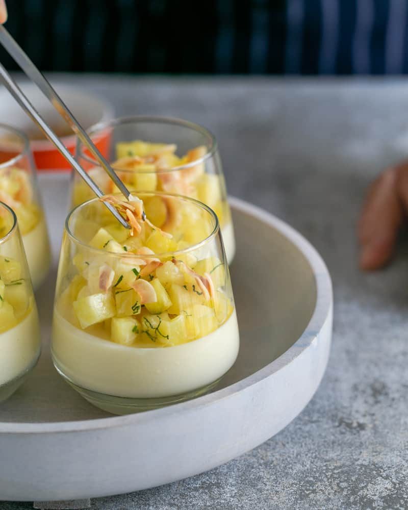 Pina colada panna cotta in a desert glass with diced pineapple, mint and toasted coconut flakes being added