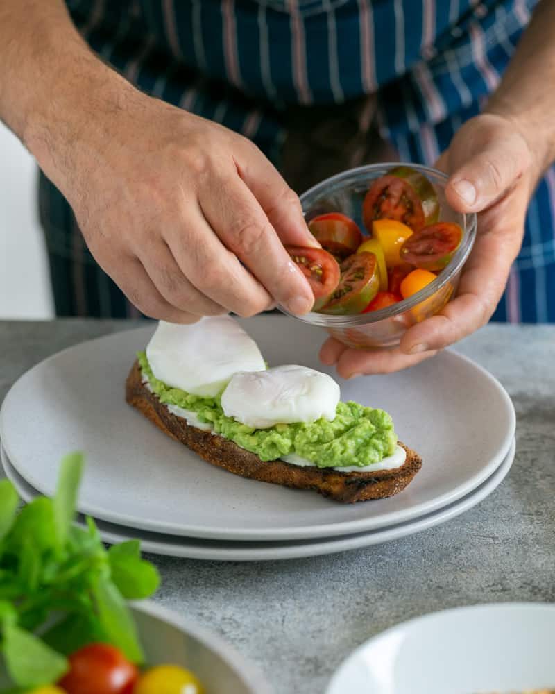 Medley of cut cherry tomatoes being placed around poached eggs on the slice of toasted sourdough bread