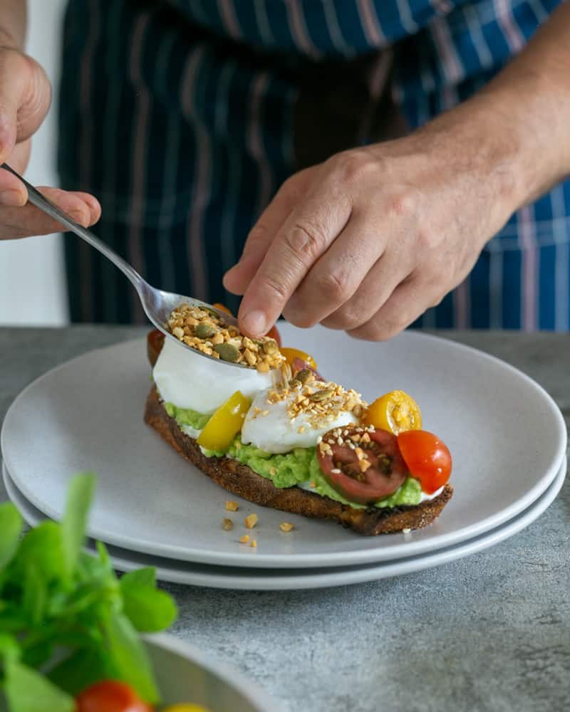 Sprinkling dukkah with a spoon on top of poached eggs and tomatoes on toasted sourdough