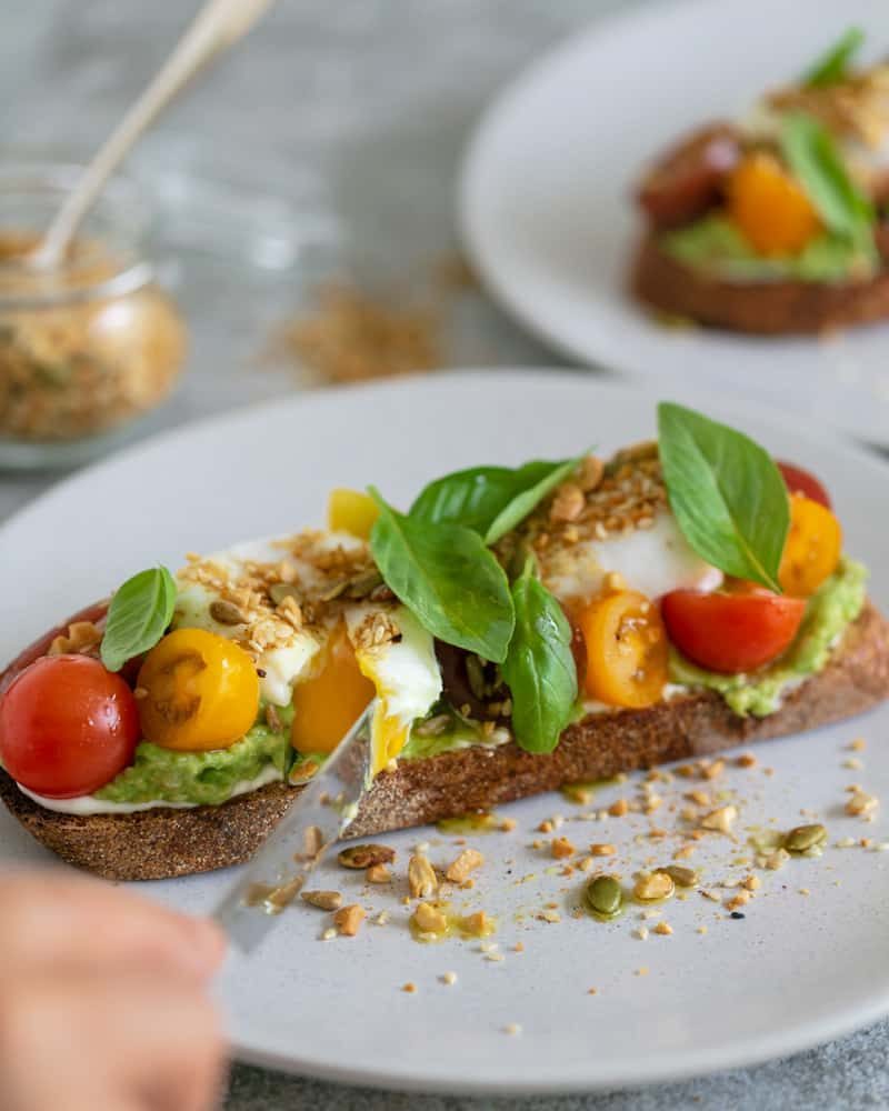 breakfast set up on table with Slice of sourdough with ricotta, crushed avocado, cut poached eggs with yolk flowing, cherry tomatoes, basil and dukkah