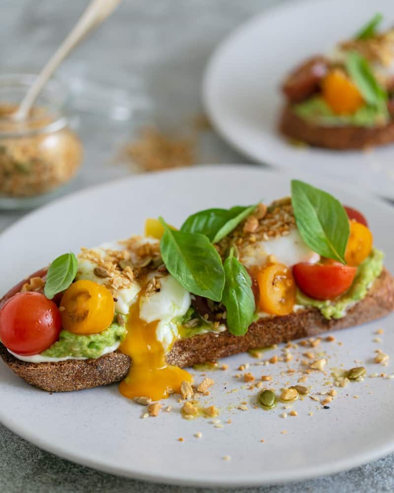 breakfast set up on table with Slice of sourdough with ricotta, crushed avocado, poached eggs cut with egg yolk oozing out, , cherry tomatoes, basil and dukkah