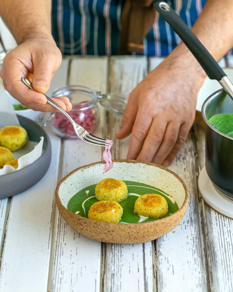 Fried paneer and potato dumplings placed in the middle of the bowl with the spinach soup, garnished with pickled onions