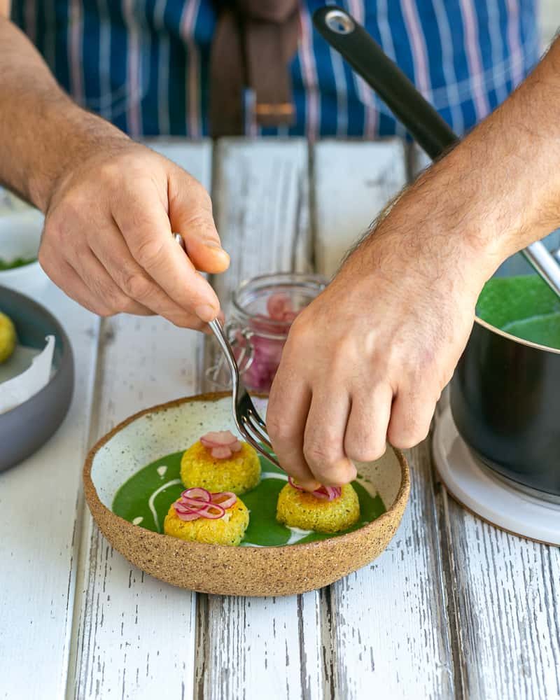 Fried paneer and potato dumplings placed in the middle of the bowl with the spinach soup, garnished with pickled onions