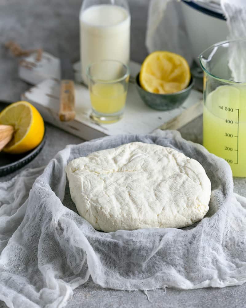 Fresh Paneer block in the muslin cloth after weight has been removed from the top with lemons, lemon juice and milk in the background showing how to make paneer