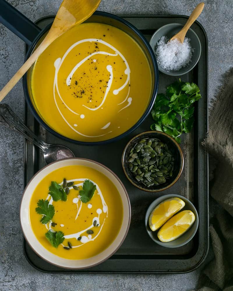 Curried pumpkin soup in a pot and a small bowl garnished with fresh coriander, pepitas and coconut milk served in a rustic metal tray